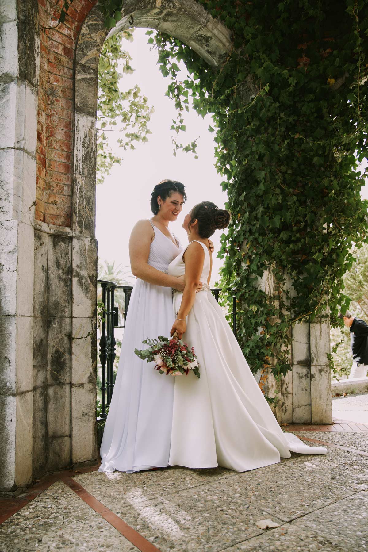 Pareja de novias posando juntas bajo un arco de piedra cubierto de vegetación durante su boda en Bizkaia