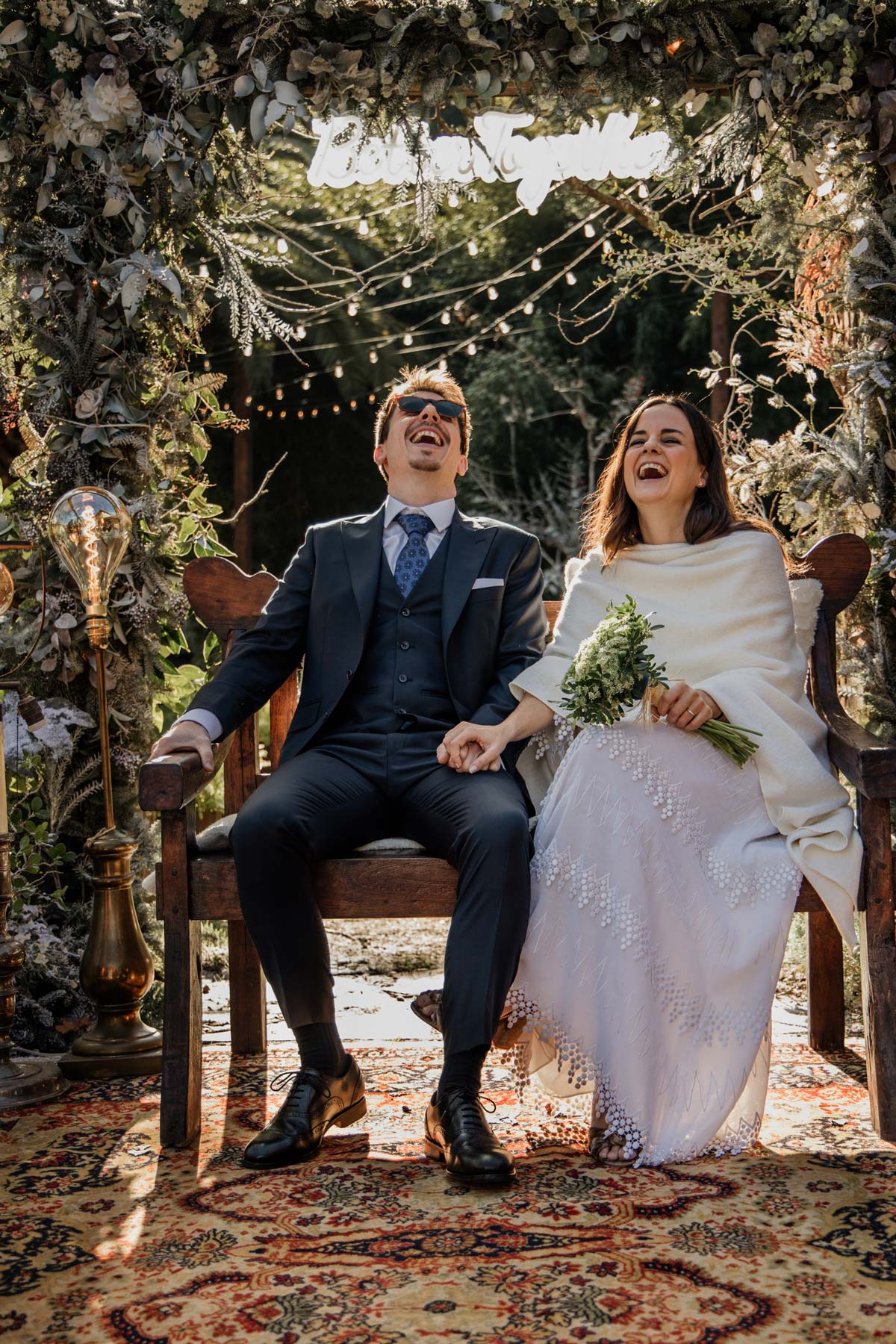 Escena de ceremonia nupcial en Bizkaia con la pareja riendo bajo un arco floral iluminado.