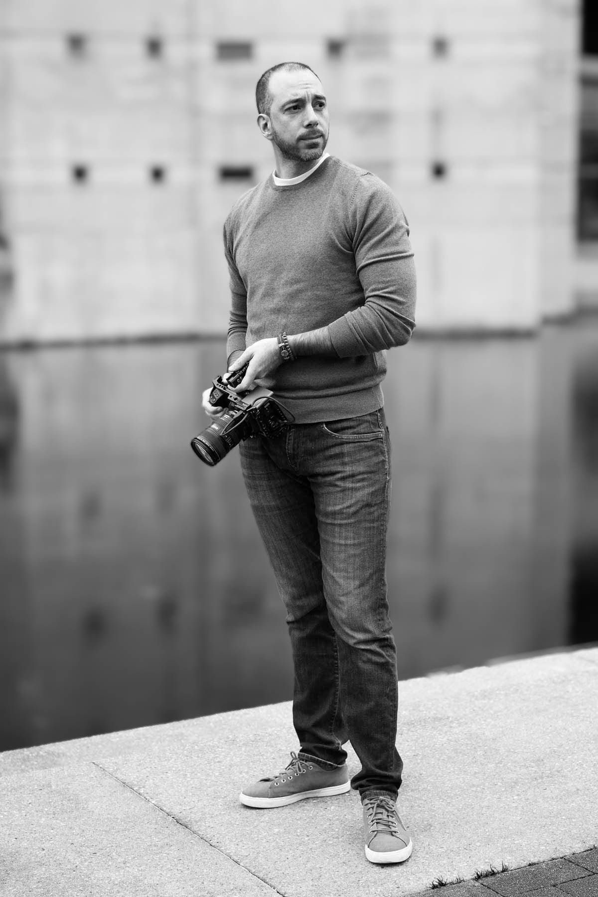 "Retrato en blanco y negro de un videógrafo sosteniendo una cámara mientras posa en exterior."
