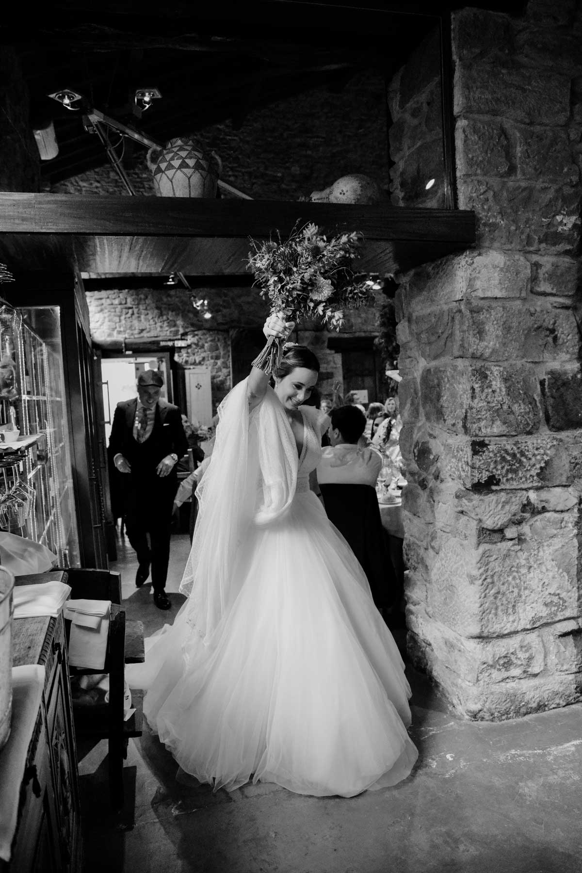 Novia levantando el ramo al entrar en un espacio rústico durante su boda, fotografía documental en Bizkaia.