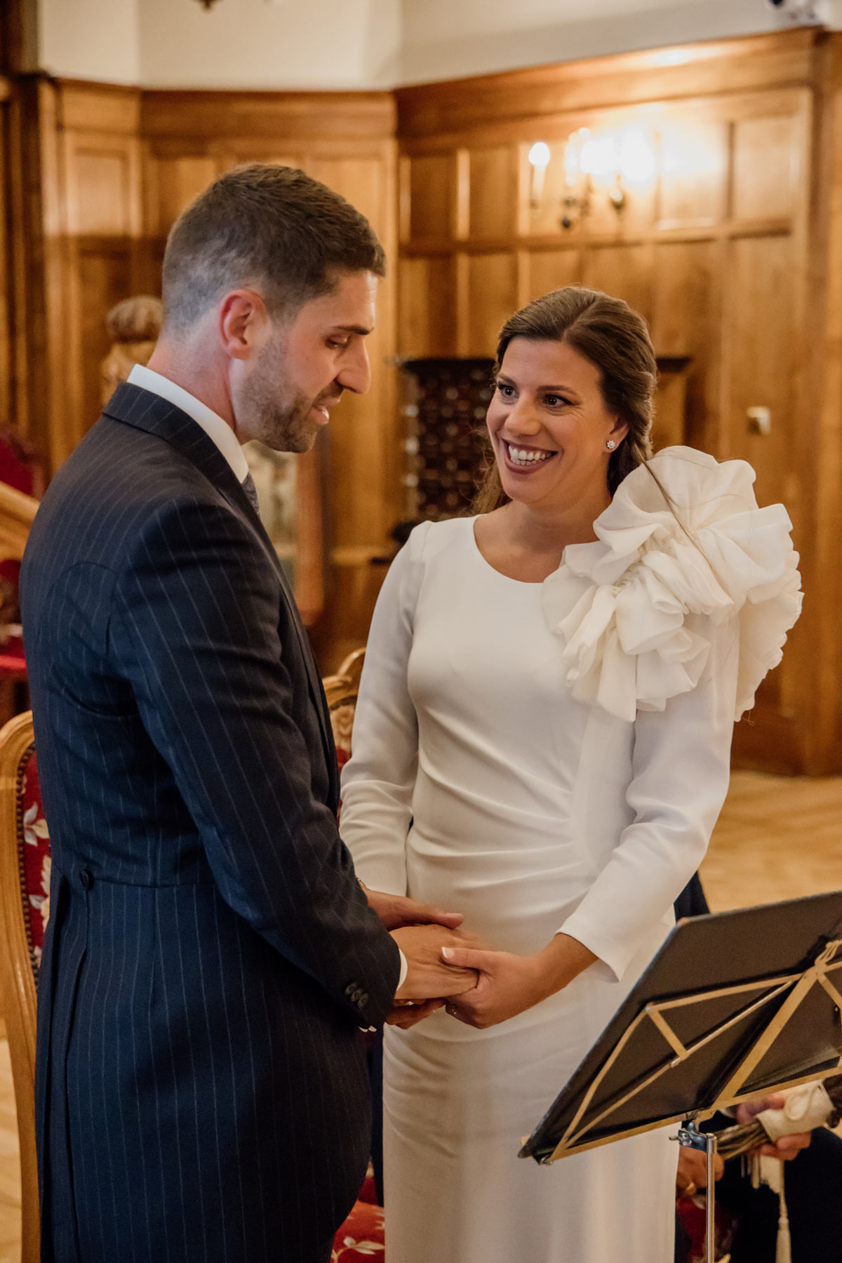 Pareja de novios sonriendo mientras se toman de las manos durante la ceremonia en un salón de madera.