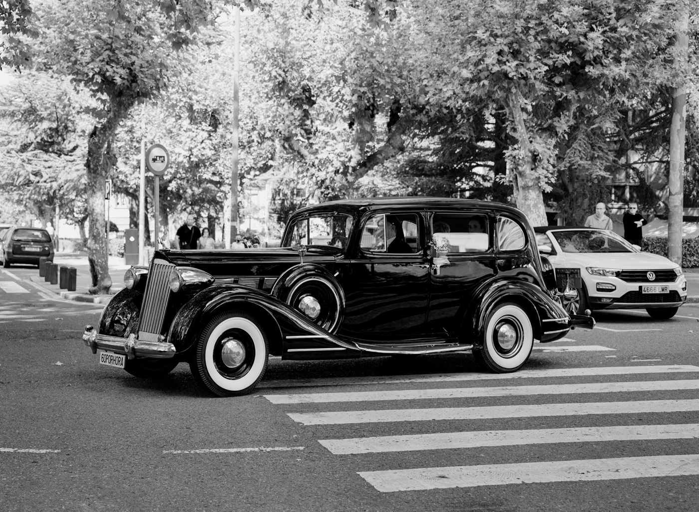 Coche clásico utilizado en una boda, fotografiado en blanco y negro mientras circula por una calle urbana.