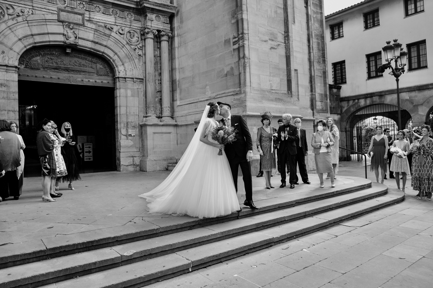 Pareja de recién casados besándose a la salida de la iglesia mientras los invitados los esperan en las escaleras.