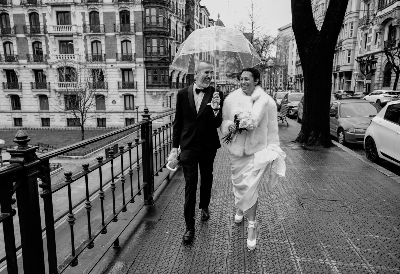 Pareja de novios caminando bajo la lluvia por una calle de Bizkaia con un paraguas transparente — fotografía natural y espontánea de North Producciones