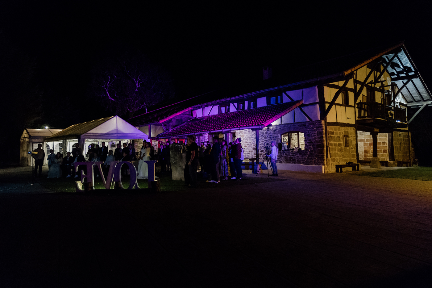 "Boda nocturna en el caserío Pagaigoikoa con iluminación de colores e invitados celebrando al aire libre en Bizkaia."