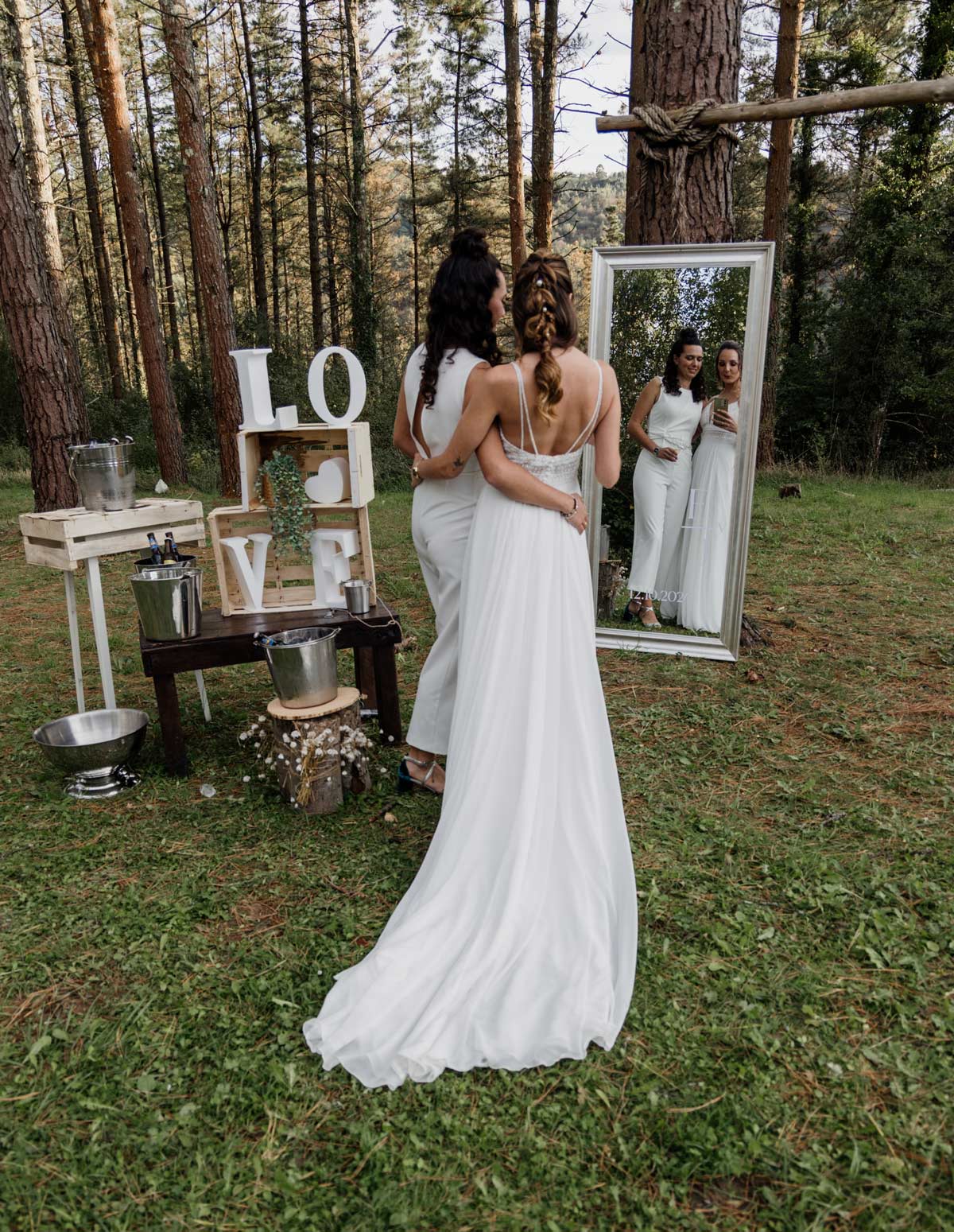 "Dos novias frente a un espejo en un rincón decorado con letras LOVE durante su boda en un bosque de Bizkaia."