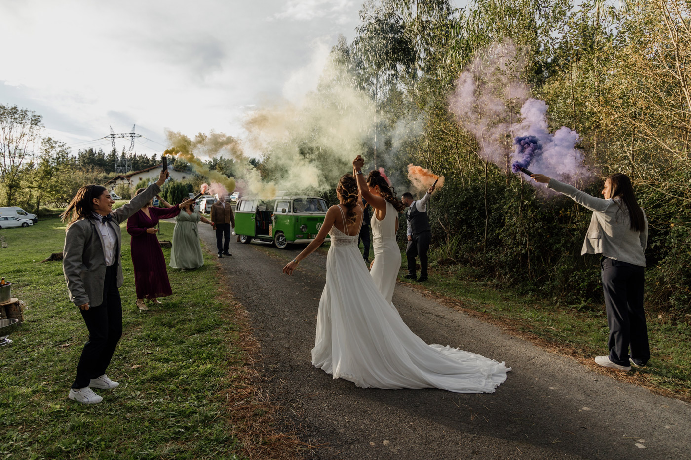 "Dos novias caminando mientras sus invitados las celebran con bengalas de colores en un camino rural, fotografía de boda en Bizkaia."