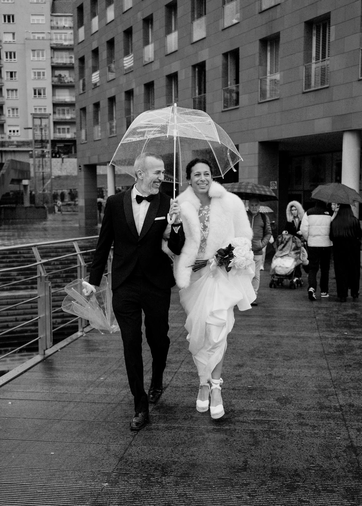 Novios caminando bajo la lluvia con un paraguas transparente en una escena natural y espontánea, fotografía realizada por un fotógrafo de bodas en Bilbao.