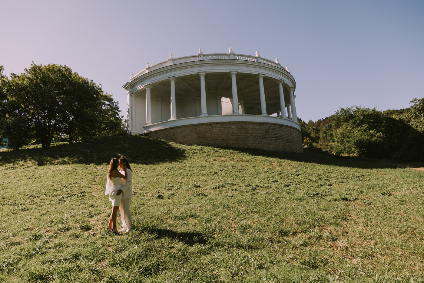 Pareja de novias abrazándose en el Torreón de Zarautz bajo un edificio circular blanco, fotografía de boda luminosa y natural.