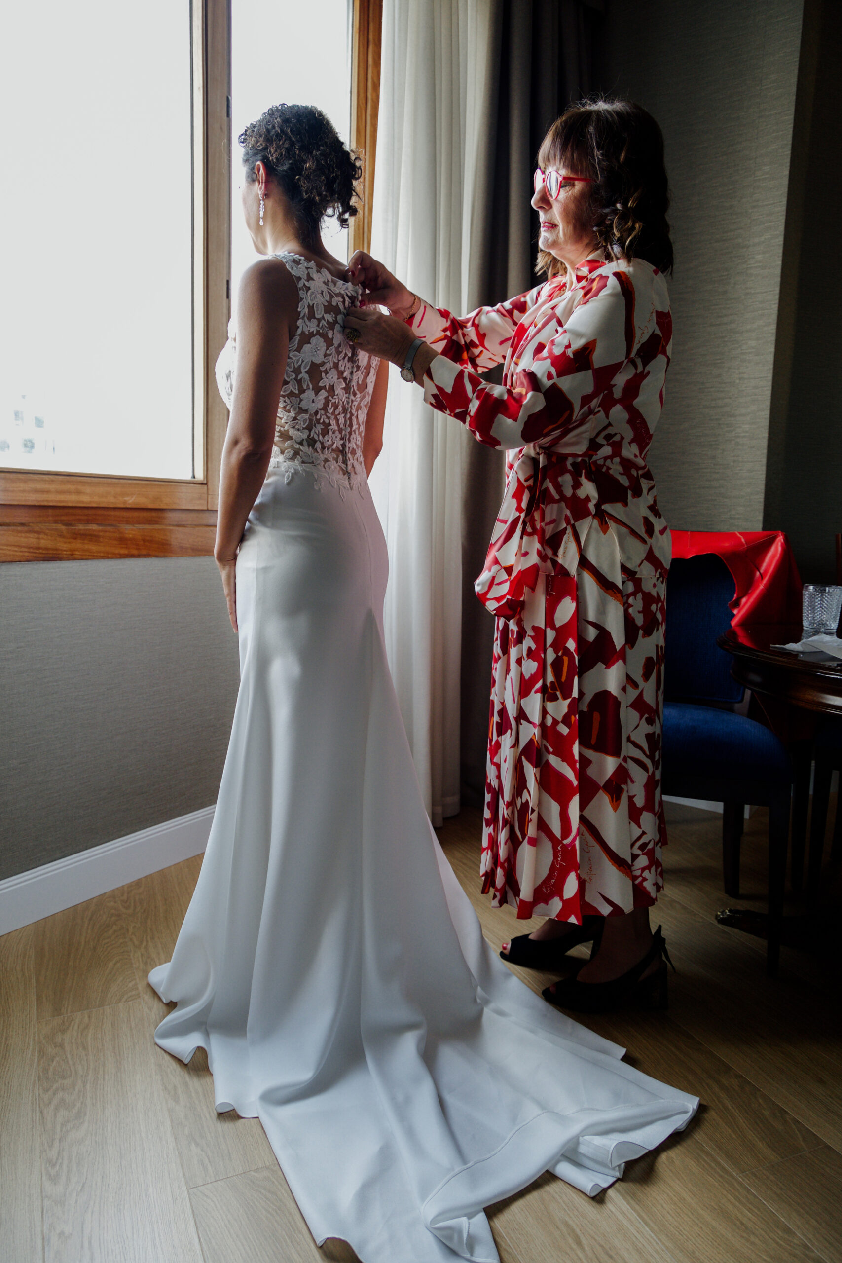 Novia preparándose para su boda mientras otra persona la ayuda con el vestido, junto a una ventana con luz natural en una habitación de Bilbao.