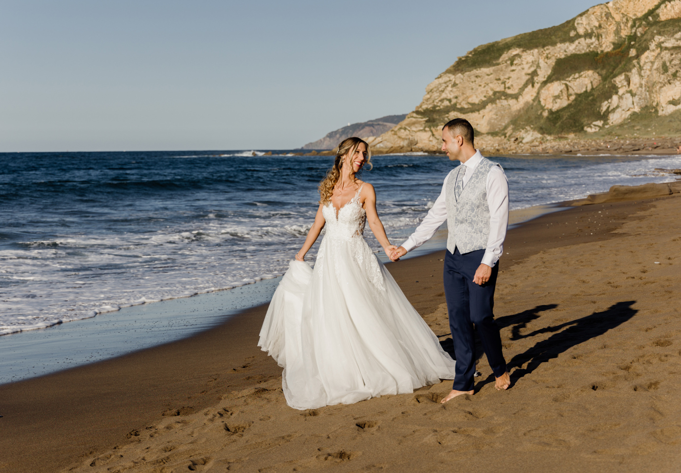 Pareja de novios caminando descalzos por la playa durante su postboda, con acantilados al fondo - Fotógrafo de bodas en Bilbao у Bizkaia.