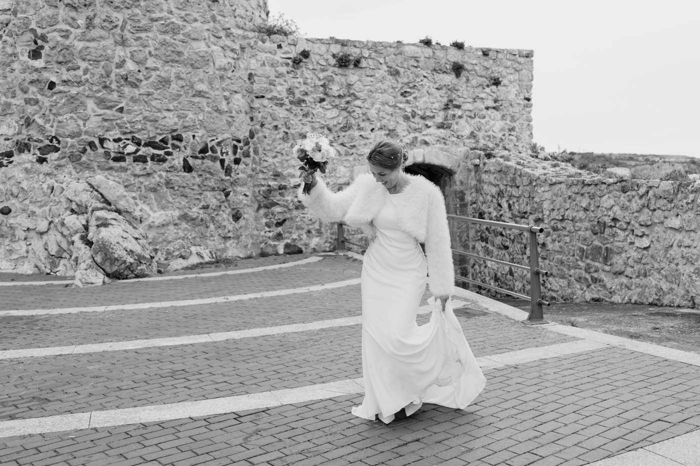 Novia caminando con su ramo en un entorno de piedra junto al mar; fotografía de boda en Cantabria en blanco y negro realizada por fotógrafo de bodas.