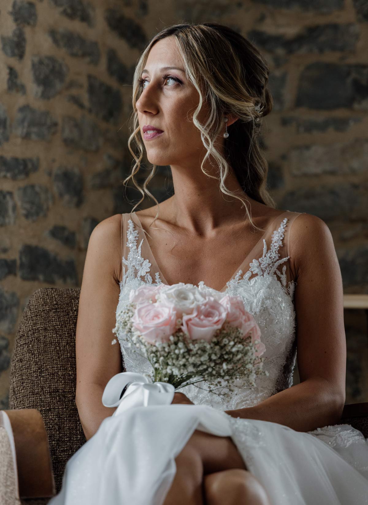 Novia sentada en interior con pared de piedra, sosteniendo su ramo y mirando hacia un lado, fotografiada por un fotógrafo de bodas en Bilbao.