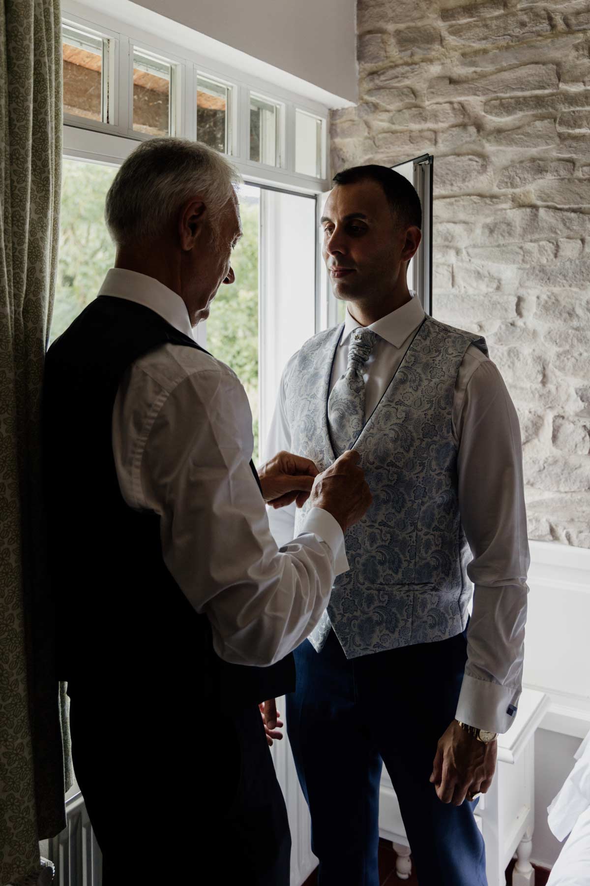 Hombre mayor ayudando al novio a ajustar el chaleco durante los preparativos de la boda, fotografiado por un fotógrafo de bodas en Bilbao.