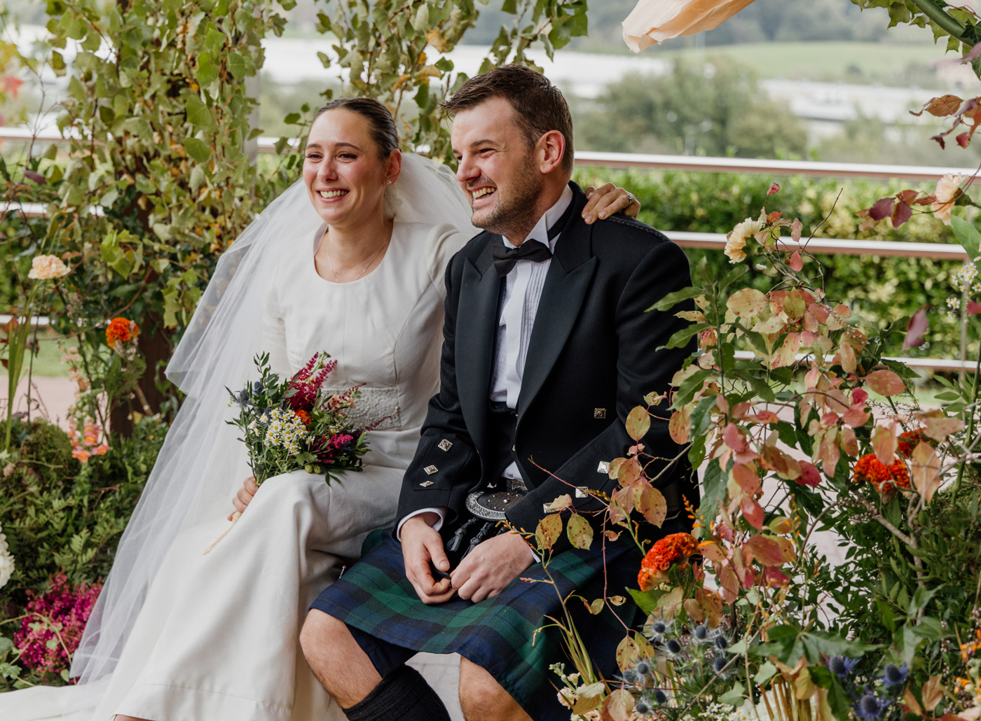 Pareja de recién casados sentada y sonriendo entre vegetación y flores, fotografía de boda al aire libre realizada por un fotógrafo de bodas en Bizkaia.