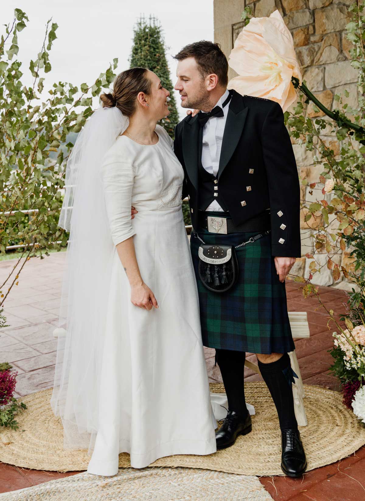 Pareja de recién casados abrazándose у mirándose con cariño durante su boda en un entorno exterior con decoración floral.