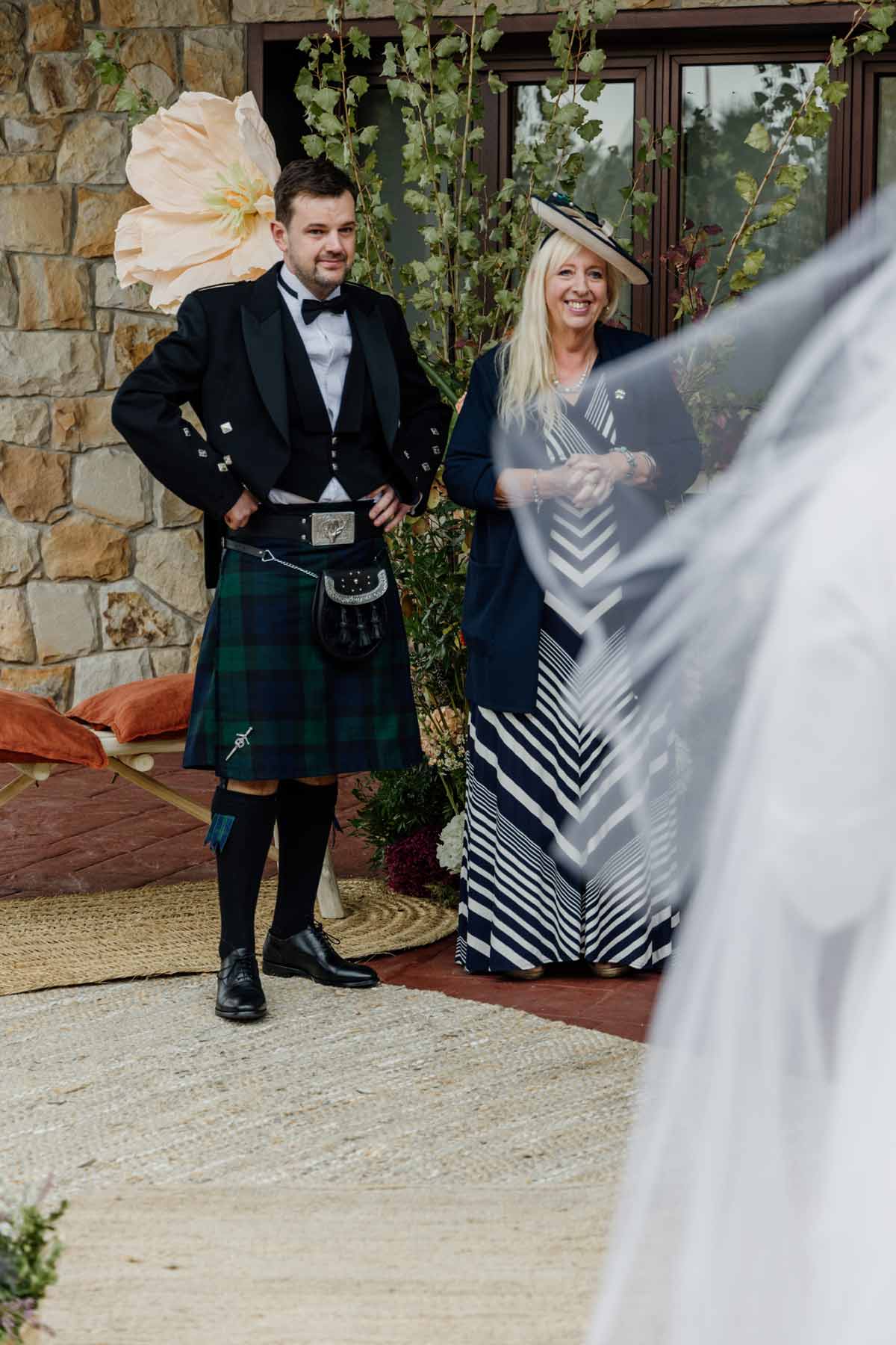 Hombre con traje tradicional y mujer sonriendo durante una boda en un entorno exterior con decoración floral.