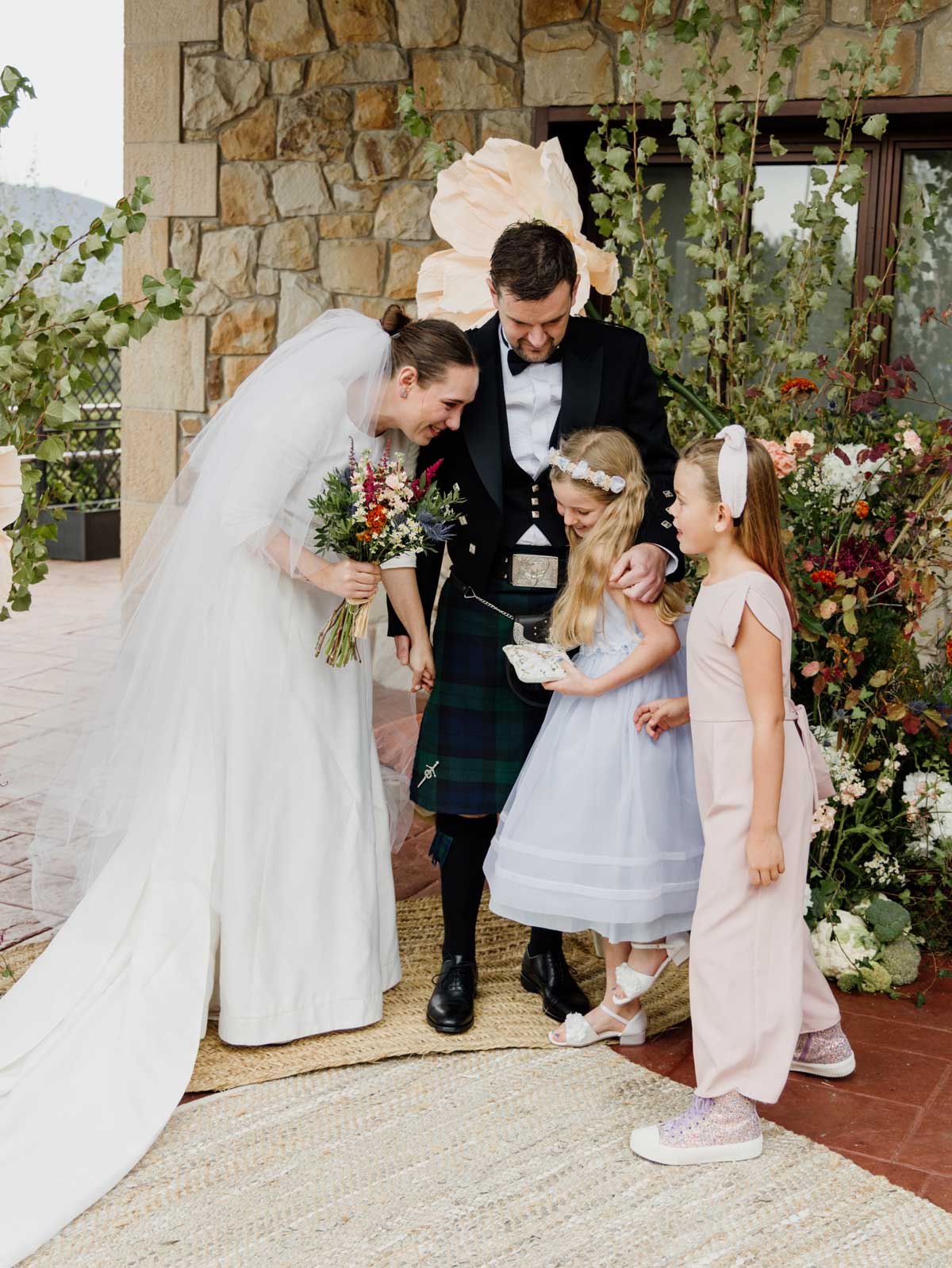 Pareja de recién casados interactuando con dos niñas durante la ceremonia en un altar decorado con flores.