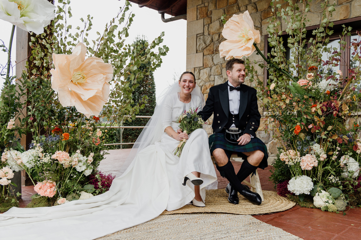 Pareja de recién casados sentada en un altar decorado con grandes flores y vegetación durante su boda.