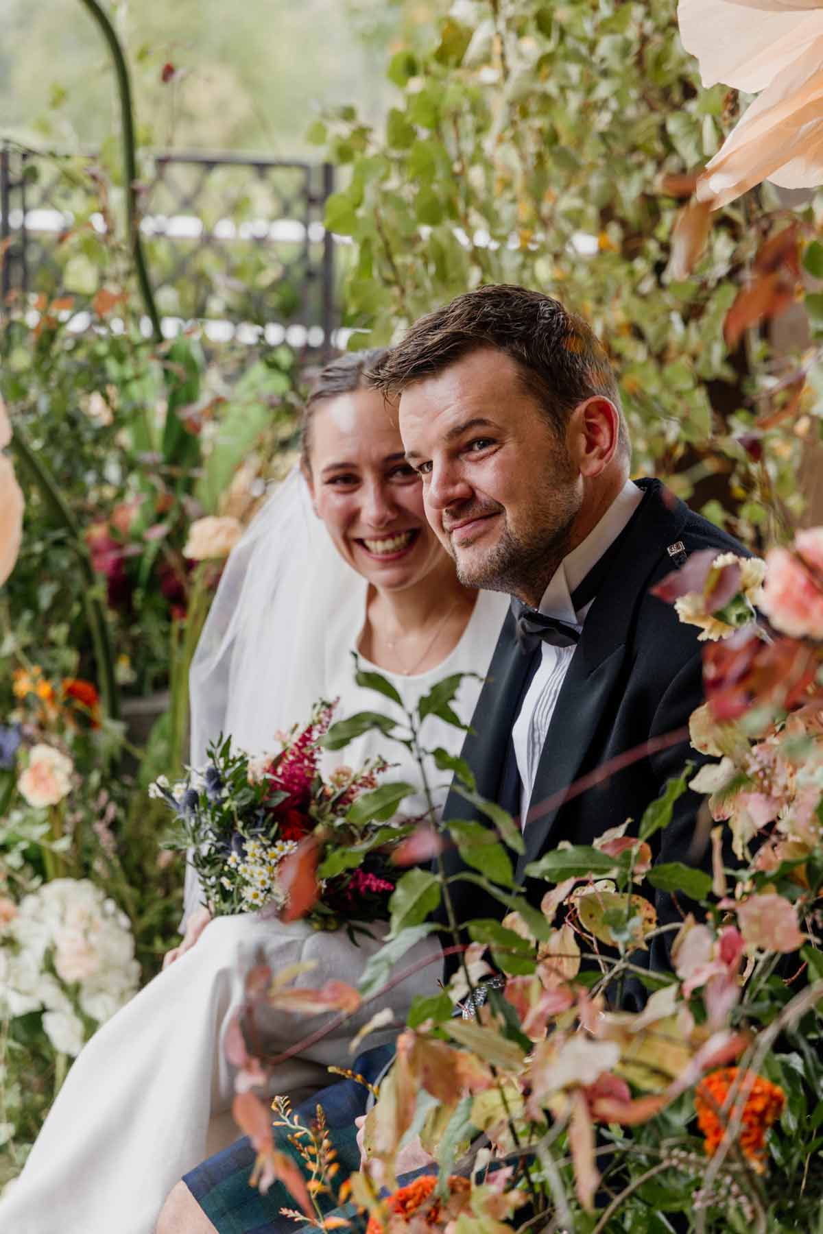 Pareja de recién casados posando entre flores y vegetación durante su boda, sonriendo y abrazados.