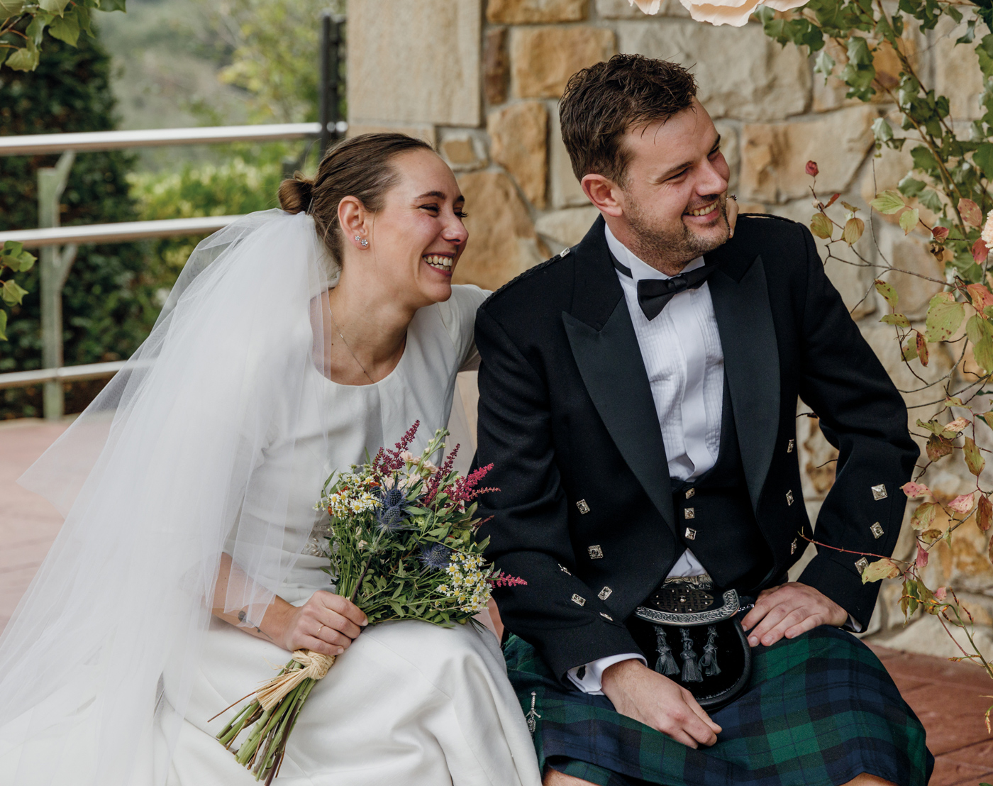 Pareja de recién casados sonriendo y sentada al aire libre durante su boda, con un ramo de flores y un fondo de pared de piedra.