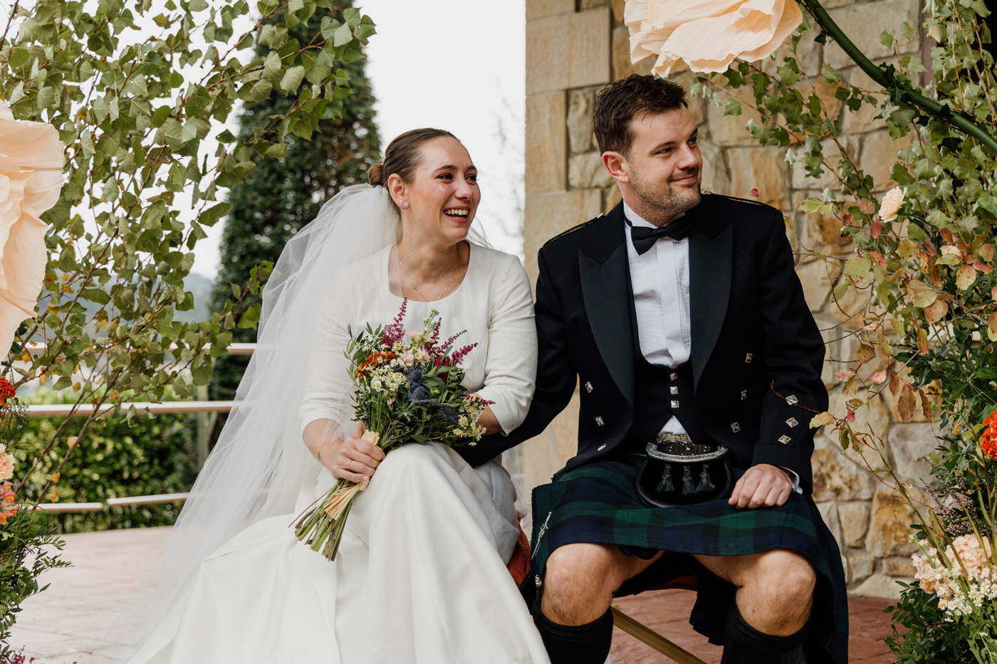 Pareja de recién casados sentados al aire libre durante la boda, sonrientes y rodeados de vegetación.