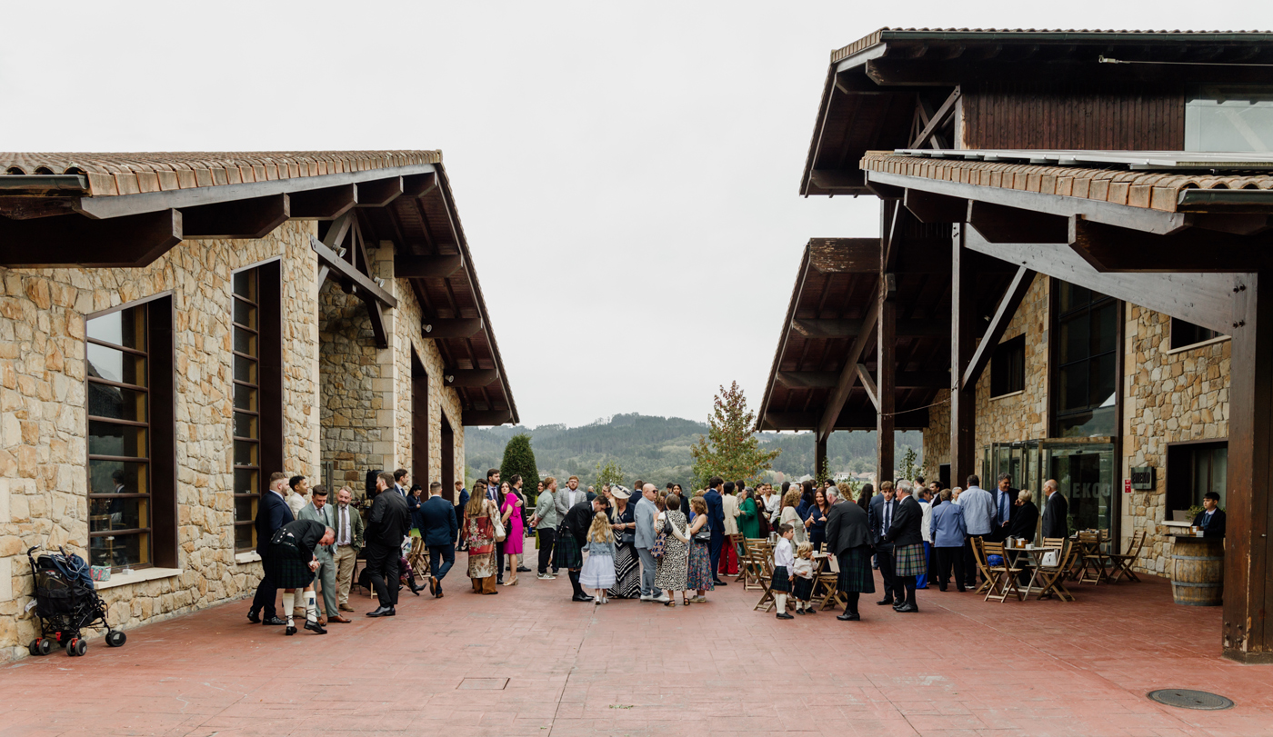 Invitados reunidos en el exterior de un restaurante rural durante una boda, con edificios de piedra y montañas al fondo