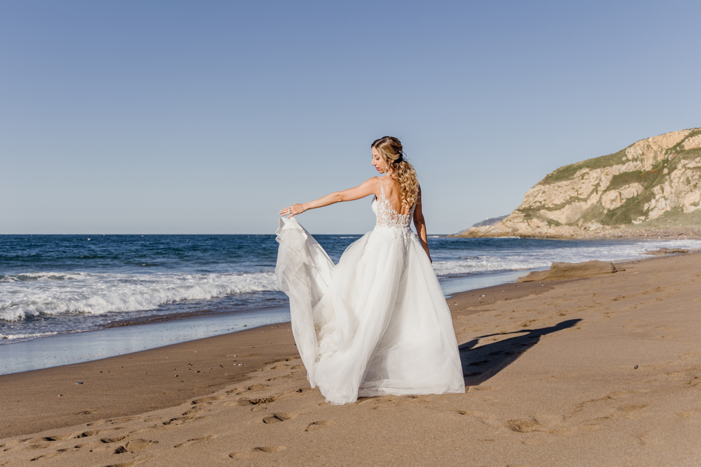 Novia caminando sobre la arena con su vestido de boda durante una sesión de postboda en una playa de Bizkaia.