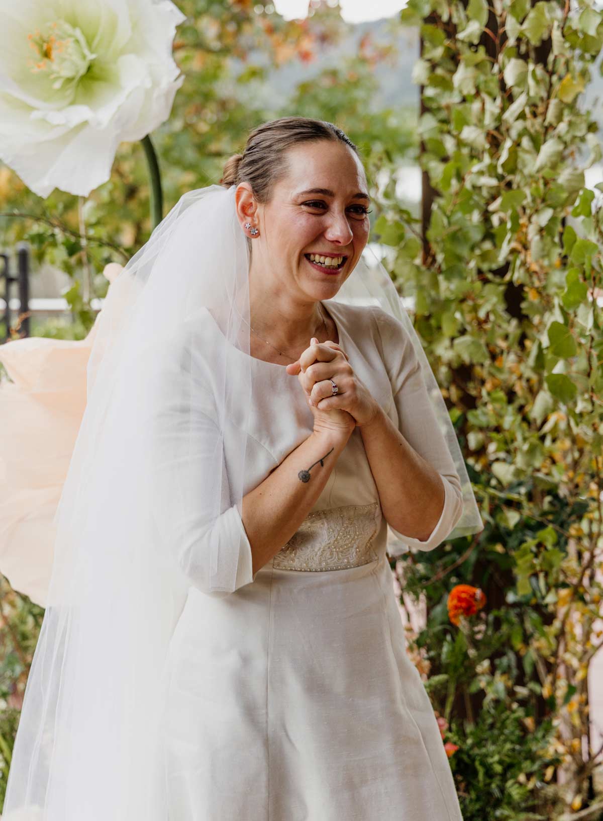 Novia emocionada sonriendo el día de su boda, vestida de blanco en un jardín con flores